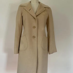 Puede incluir: Un abrigo color beige con un diseño clásico. El abrigo presenta un cuello con solapas, dos bolsillos delanteros grandes y dos botones para cerrar. El abrigo se exhibe en un maniquí.