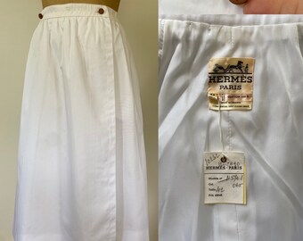 May include: White cotton wrap skirt with a brown button closure and a label that reads "Hermès Paris" and "Cotton 100% Made in France".