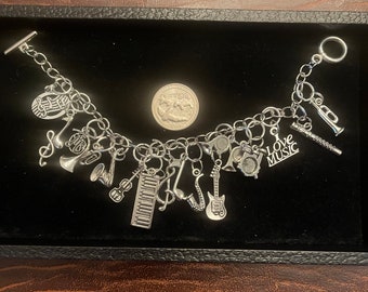 May include: Silver charm bracelet featuring musical instrument charms, including a trumpet, guitar, piano keys, and saxophone. The bracelet also has charms with musical notes and the words "Love Music". The bracelet is displayed on a black surface.