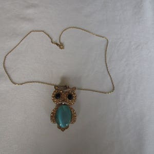Pu&ograve; includere: Una collana con catena dorata con un ciondolo a forma di gufo. Il gufo ha una pietra preziosa blu turchese al centro e occhi neri.