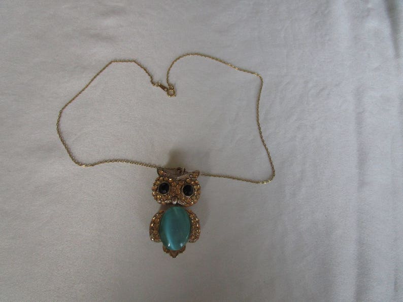 Pu&ograve; includere: Una collana con catena dorata con un ciondolo a forma di gufo. Il gufo ha una pietra preziosa blu turchese al centro e occhi neri.