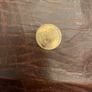 Puede incluir: Una moneda de color dorado con un diseño que presenta un patrón floral en el centro.