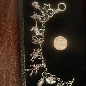 May include: A silver-toned charm bracelet featuring various dangling charms including a guitar, a star, an American flag, a bird, the letters OH, an airplane, an astronaut, a football, an Ohio state shape, and the letters USA, next to a US quarter.
