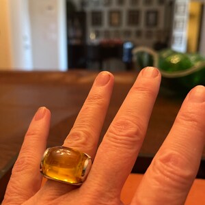 May include: A large, amber-colored ring with a rectangular, rounded design. The ring features a thick, clear, amber-colored stone set in a silver-colored metal band. The ring is worn on a finger.