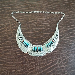 May include: A silver-toned necklace with a crescent-shaped pendant. The pendant features inlaid turquoise, black, and red stones, with intricate leaf and feather designs. The necklace has a chain closure.