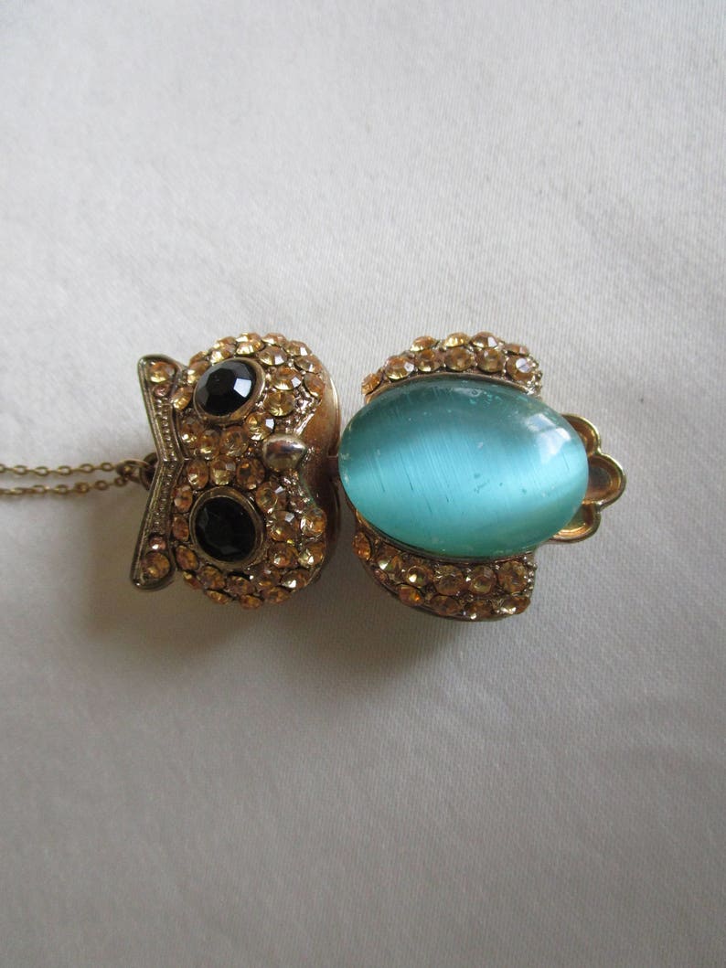 Pu&ograve; includere: Una collana con ciondolo a forma di gufo in oro, decorata con una pietra preziosa blu e strass trasparenti. Il gufo ha gli occhi neri ed &egrave; circondato da strass.