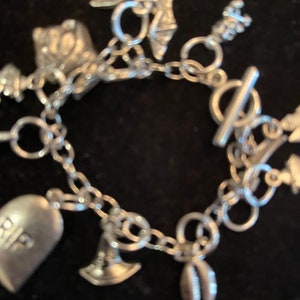 Könnte beinhalten: Ein silbernes Charm-Armband mit verschiedenen Charms, darunter ein Grabstein mit der Aufschrift "RIP", eine Muschel, ein Anker und ein kleines Haus.