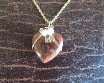 May include: A heart-shaped pendant necklace. The pendant is made of a reddish-brown stone with a gold-colored wire wrap and a gold-colored chain. The pendant has a small white bead and gold wire accents.