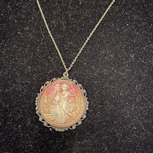Può includere: Una collana a catena d'argento con un ciondolo a cammeo raffigurante una donna che versa acqua da una brocca. Il cammeo è incastonato in una cornice d'argento con un bordo decorativo di piccole perle d'argento.