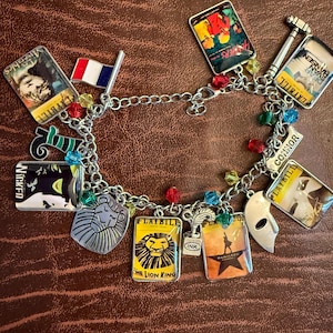 Puede incluir: Una pulsera de dijes plateada con portadas rectangulares en miniatura de programas de musicales como The Lion King, Hamilton, Wicked y Dear Evan Hansen, junto a dijes de una bandera francesa, una máscara, un león y el nombre Connor.