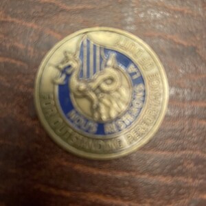 Puede incluir: Una moneda redonda de color dorado con un diseño azul y plateado. La moneda presenta una criatura similar a un dragón con alas y cola. El texto alrededor del borde de la moneda dice "Awarded for Outstanding Performance".