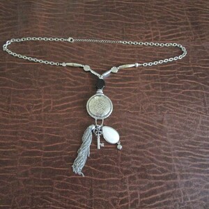 May include: A silver chain necklace with a silver pendant featuring a circular design, a key, and a white stone. The necklace also has black and silver beads.