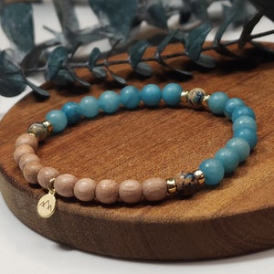 Op de afbeelding: Een kralenarmband met lichtblauwe en bruine kralen, afgewisseld met goudkleurige accenten. Een kleine gouden bedel met een berglogo hangt eraan. De armband ligt op een houten oppervlak.