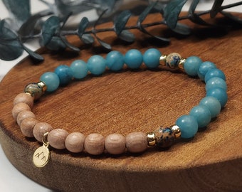 Puede incluir: Una pulsera de cuentas con cuentas azul claro y marrón, intercaladas con detalles dorados. Un pequeño dije dorado con un logotipo de montaña cuelga de la pulsera. La pulsera se muestra sobre una superficie de madera.
