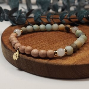May include: A beaded bracelet featuring a mix of light brown wooden beads, light green and beige gemstones, and gold accents. A small gold charm with a mountain design hangs from the bracelet. The bracelet is displayed on a wooden surface.