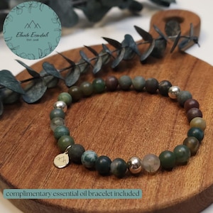 Op de afbeelding: Een armband met groene, bruine en zilveren kralen, gepresenteerd op een houten oppervlak. De armband heeft een kleine gouden bedel. De tekst "Elevate Essentials EST. 2018" staat in een cirkel linksboven. De tekst "complimentary essential oil bracelet included" staat onderaan.
