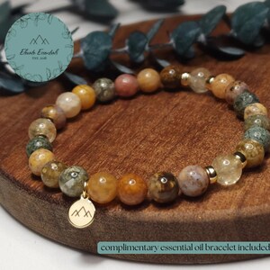 Könnte beinhalten: Ein Perlenarmband mit verschiedenen erdfarbenen Edelsteinen, darunter gelbe, braune und grüne Perlen. Ein goldfarbener Anhänger mit einem Bergdesign hängt am Armband. Das Armband wird auf einer Holzoberfläche präsentiert.