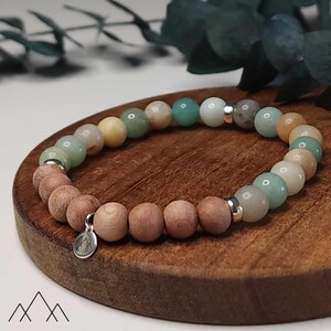 Op de afbeelding: Een kralenarmband met een mix van lichtgekleurde edelstenen en houten kralen. De armband bevat een verscheidenheid aan kleuren, waaronder groen, geel en beige, met zilverkleurige afstandhouders en een kleine bedel. De armband wordt gepresenteerd op een houten oppervlak.