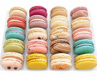 May include: A clear plastic tray containing twenty assorted French macarons arranged in four rows of five. The colorful cookies feature various pastel and vibrant shades including yellow, pink, blue, green, brown, and cream, each with a visible filling.