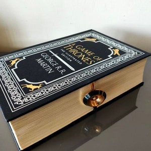 Puede incluir: Un libro de tapa dura negro con letras doradas y un borde decorativo. El t&iacute;tulo es "A Game of Thrones" y el autor es "George R.R. Martin". El libro es la edici&oacute;n ilustrada.