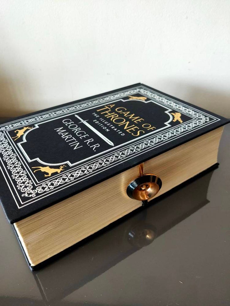Puede incluir: Un libro de tapa dura negro con letras doradas y un borde decorativo. El t&iacute;tulo es "A Game of Thrones" y el autor es "George R.R. Martin". El libro es la edici&oacute;n ilustrada.