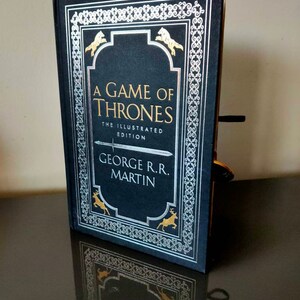Puede incluir: Un libro negro de tapa dura con un dise&ntilde;o plateado y dorado. El t&iacute;tulo "A Game of Thrones" est&aacute; impreso en dorado en la portada. El libro es la edici&oacute;n ilustrada de George R.R. Martin.