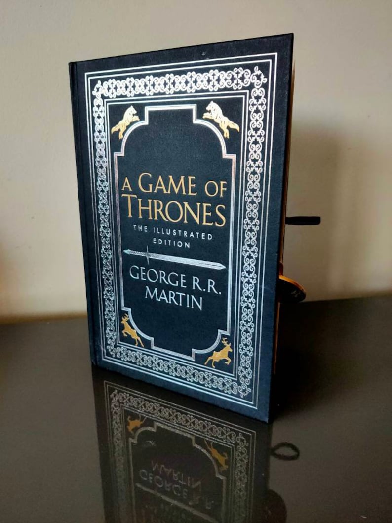 Puede incluir: Un libro negro de tapa dura con un dise&ntilde;o plateado y dorado. El t&iacute;tulo "A Game of Thrones" est&aacute; impreso en dorado en la portada. El libro es la edici&oacute;n ilustrada de George R.R. Martin.