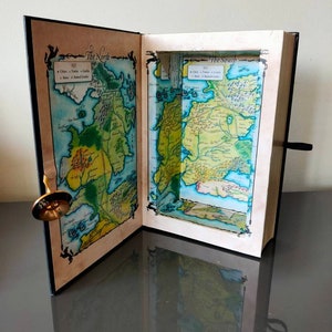 Puede incluir: Un libro negro con un compartimento oculto que se abre para revelar un mapa de Poniente de la popular serie de televisi&oacute;n Juego de Tronos. El mapa est&aacute; dividido en dos secciones, "El Norte" y "El Sur".