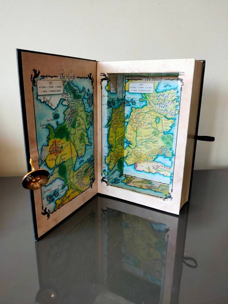 Puede incluir: Un libro negro con un compartimento oculto que se abre para revelar un mapa de Poniente de la popular serie de televisi&oacute;n Juego de Tronos. El mapa est&aacute; dividido en dos secciones, "El Norte" y "El Sur".