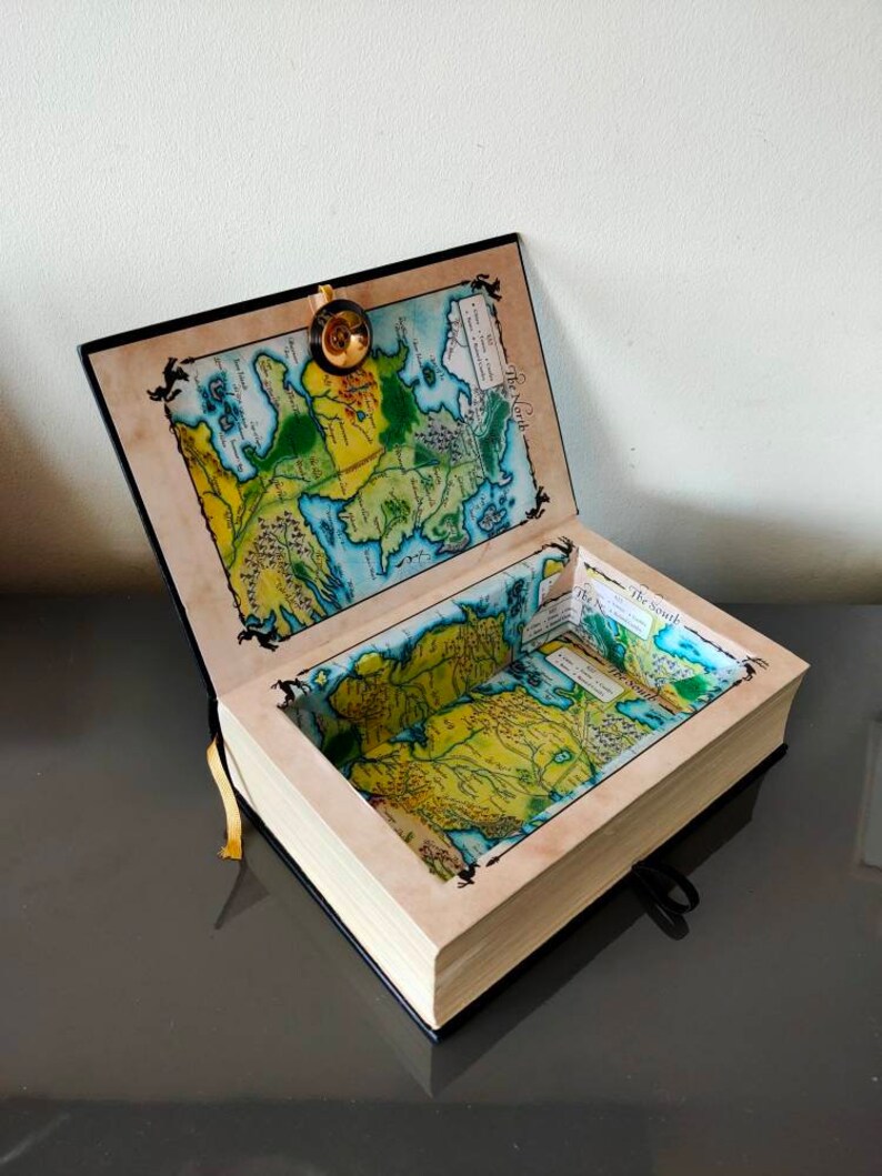 Puede incluir: Una caja negra con forma de libro con un mapa de Poniente en la portada y en el interior. La caja est&aacute; abierta y el mapa es visible en el interior. La caja tiene un cierre dorado y una cinta negra.