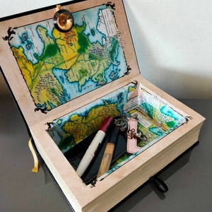 Puede incluir: Un libro negro con un mapa de Poniente en la cubierta interior. El libro est&aacute; abierto y tiene un peque&ntilde;o compartimento en el interior. El compartimento contiene un llavero, un l&aacute;piz labial y una brocha de maquillaje.