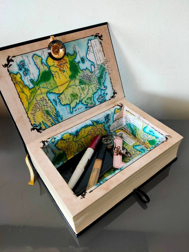 Puede incluir: Un libro negro con un mapa de Poniente en la cubierta interior. El libro est&aacute; abierto y tiene un peque&ntilde;o compartimento en el interior. El compartimento contiene un llavero, un l&aacute;piz labial y una brocha de maquillaje.