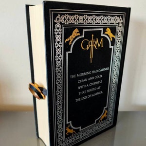 Puede incluir: Un libro de tapa dura negro con un dise&ntilde;o dorado y plateado. El t&iacute;tulo del libro es "GARM" y el texto de la portada dice "The morning had dawned clear and cold, with a crispness that hinted at the end of summer."