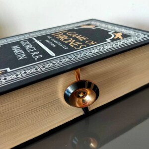 Puede incluir: Un libro negro de tapa dura con letras doradas titulado "A Game of Thrones" de George R.R. Martin. El libro es la edici&oacute;n ilustrada y tiene un marcap&aacute;ginas dorado.