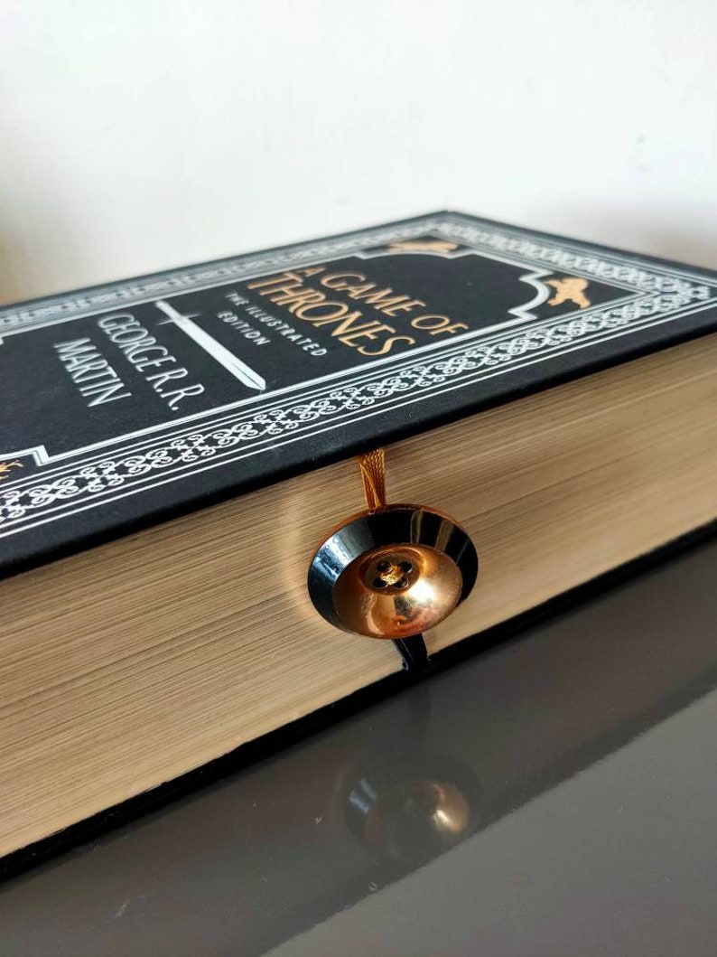 Puede incluir: Un libro negro de tapa dura con letras doradas titulado "A Game of Thrones" de George R.R. Martin. El libro es la edici&oacute;n ilustrada y tiene un marcap&aacute;ginas dorado.