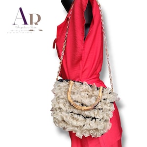 May include: A cream-colored handbag with a bamboo handle and a chain strap. The bag is adorned with ruffled fabric and has a black interior. The image also displays the text "AR Angelie Rose ACCESSORIES FOR DONNA."