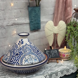 May include: A blue and white ceramic tagine with intricate floral designs. The tagine is on a white lace tablecloth with a small chalkboard sign that says "$8.99".