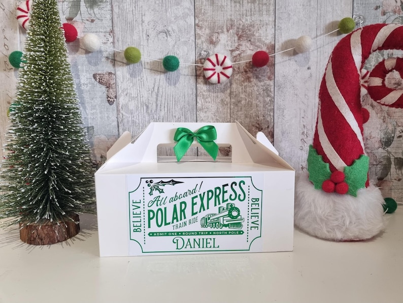 May include: A white gift box with a green bow on top, featuring a green and white "Polar Express" ticket design. The box has a handle and is decorated with a small Christmas tree and a candy cane-shaped decoration.