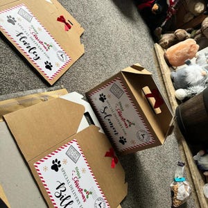 Puede incluir: Cajas de cart&oacute;n con etiquetas navide&ntilde;as que dicen "Entrega especial para:" seguidas de nombres. Las etiquetas presentan huellas de patas, copos de nieve y "Feliz Navidad". Algunas cajas est&aacute;n adornadas con lazos rojos.