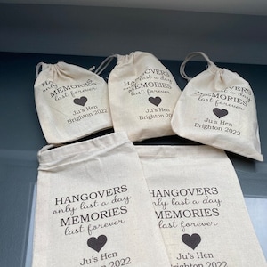 May include: Five white cotton drawstring bags with black text that reads "Hangover's only last a day Memories last forever" and "Ju's Hen Brighton 2022".
