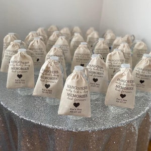 May include: A group of small, white fabric bags with brown drawstring closures. Each bag contains a clear plastic water bottle and has a brown printed design that reads "Hangovers only last a day, Memories last forever." The bags are arranged on a silver sparkly table cloth.
