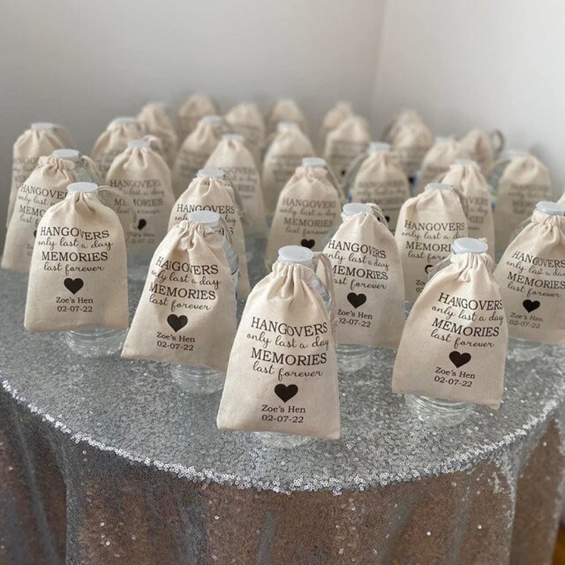 May include: A group of small, white fabric bags with brown drawstring closures. Each bag contains a clear plastic water bottle and has a brown printed design that reads "Hangovers only last a day, Memories last forever." The bags are arranged on a silver sparkly table cloth.