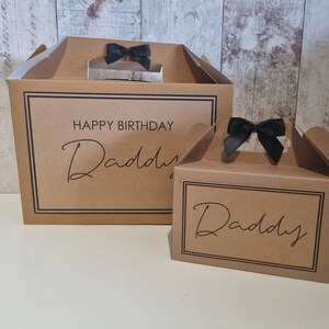May include: Two brown cardboard gift boxes with black ribbon handles. The larger box has the text "Happy Birthday Daddy" printed on the front. The smaller box has the text "Daddy" printed on the front.