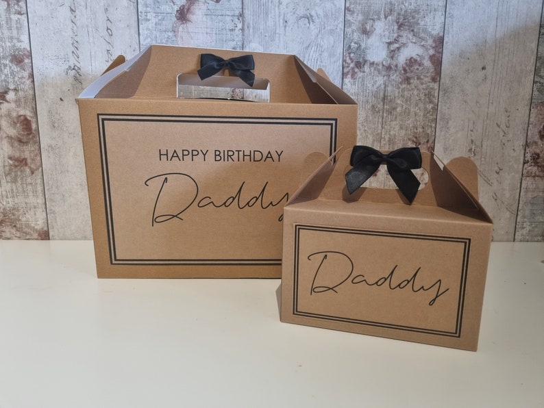 May include: Two brown cardboard gift boxes with black ribbon handles. The larger box has the text "Happy Birthday Daddy" printed on the front. The smaller box has the text "Daddy" printed on the front.