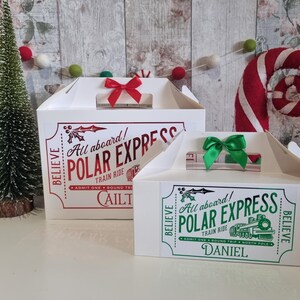 May include: Two white cardboard gift boxes with handles, decorated for Christmas. The larger box has a red bow and text that reads "All aboard! POLAR EXPRESS TRAIN RIDE." The smaller box has a green bow and the text "All aboard! POLAR EXPRESS TRAIN RIDE."