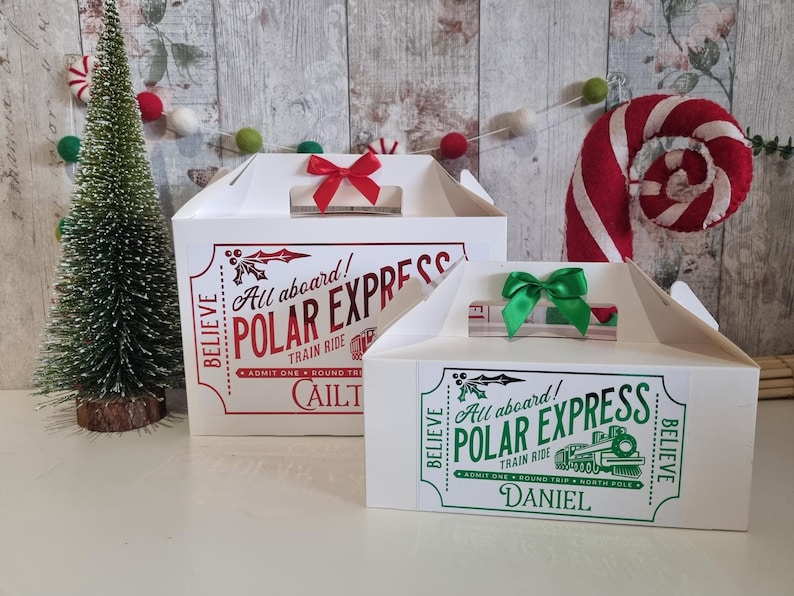May include: Two white cardboard gift boxes with handles, decorated for Christmas. The larger box has a red bow and text that reads "All aboard! POLAR EXPRESS TRAIN RIDE." The smaller box has a green bow and the text "All aboard! POLAR EXPRESS TRAIN RIDE."