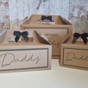 May include: Three brown cardboard gift boxes with black ribbon bows and the word "Daddy" printed on them. The largest box has the words "Happy Birthday" printed on it.
