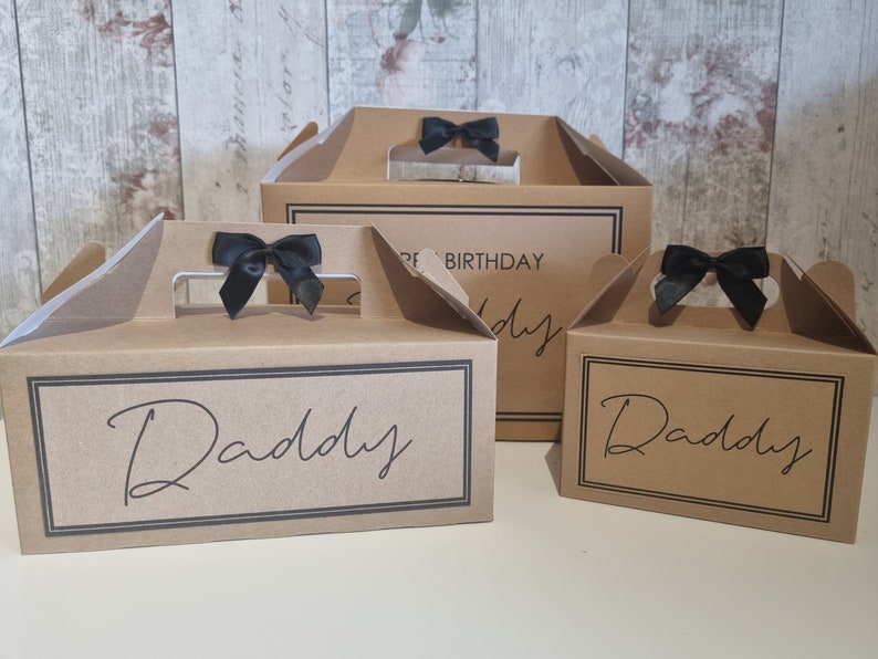 May include: Three brown cardboard gift boxes with black ribbon bows and the word "Daddy" printed on them. The largest box has the words "Happy Birthday" printed on it.