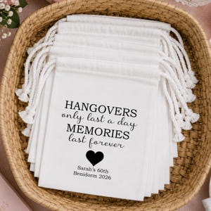 May include: A stack of white fabric drawstring bags with black text that reads HANGOVERS only last a day MEMORIES last forever, a black heart icon, and the text Sarah's 60th BENIDORM 2026. Each bag features white braided cotton drawstrings.