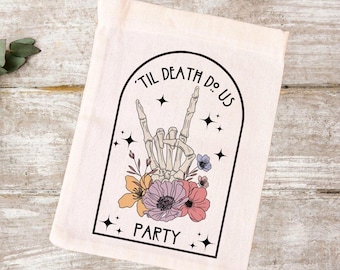 May include: A white cotton tote bag with a black graphic design. The graphic features a skeleton hand making a rock and roll hand gesture with flowers around it. The text "'Til Death Do Us Party" is written in a black arch above the hand.